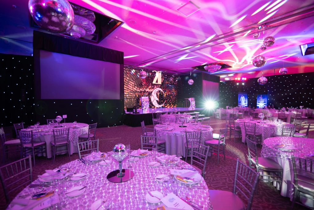 Elegant dinner setup in a lavish event hall with ambient lighting and disco balls.